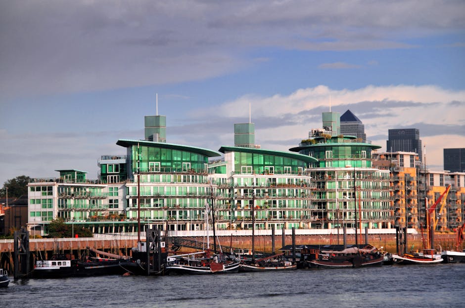 A modern, multi-storey residential building with a distinctive curved roof design and extensive exterior green glass windows, situated along a waterfront. The building features several terraces and ba