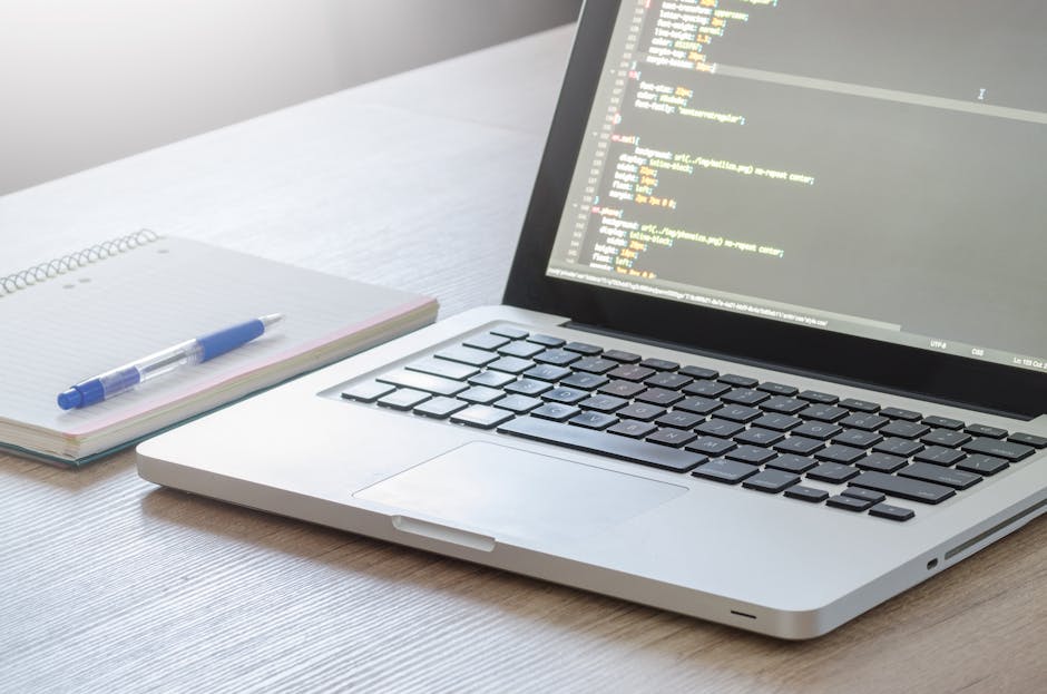 A close-up image of a workspace showing a silver laptop displaying lines of code on its screen, placed on a light wooden desk. To the left of the laptop, there is an open spiral-bound notebook with a 