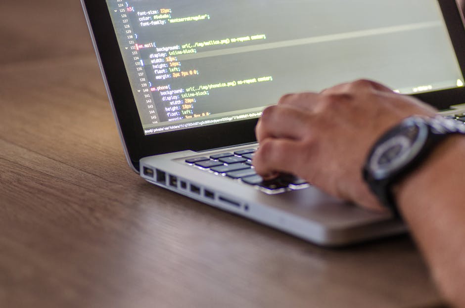 A close-up image of a workspace showing a silver laptop displaying lines of code on its screen, placed on a light wooden desk. To the left of the laptop, there is an open spiral-bound notebook with a 